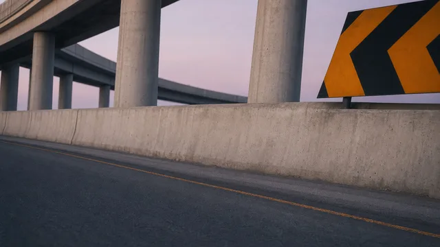 An empty road beneath concrete flyovers with barriers and a yellow-and-black turn sign; a setting reference.